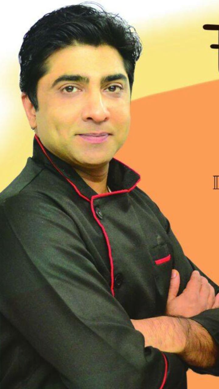 Portrait of a professional chocolatier wearing a black chef's coat with red trim, smiling with arms crossed against a warm background.